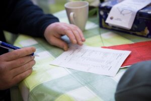 A volunteer goes through a packing list form with a recipient. Only the volunteer’s hands are in shot, and they are holding a blue pen, with a mug next to them on the table.
