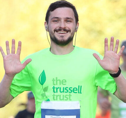 A fundraiser takes part in the Royal Parks Half Marathon. They have dark brown hair and a beard, and they are wearing a bright green Trussell Trust t-shirt with their race number attached. They are smiling into the camera and waving both hands as they run.