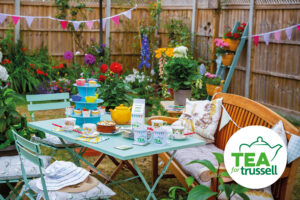 A garden table with a cake stand, mugs and a teapot. There are chairs and a bench around the table, with flowers and bunting in the background.