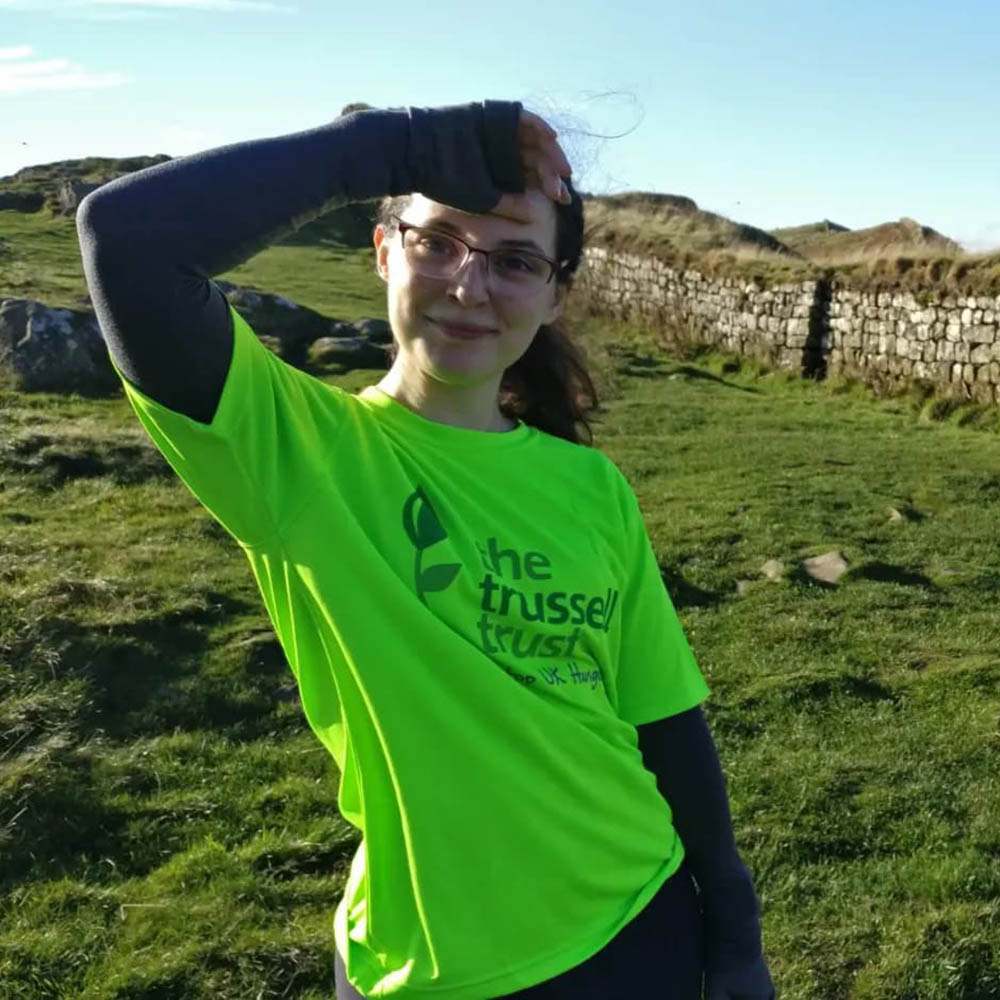 A fundraiser stands next to Hadrian’s Wall while taking part in the Lord of the Rings Challenge. They have long brown hair tied back, and are wearing glasses and a bright green Trussell Trust t-shirt. They are smiling into the camera and raising a hand above their head to keep the sun out of their eyes.