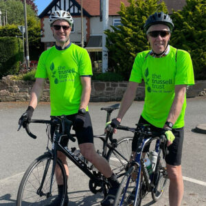 Two fundraisers smile into the camera as they stand with their bikes. Both are wearing bright green Trussell Trust t-shirts, safety helmets and sunglasses.