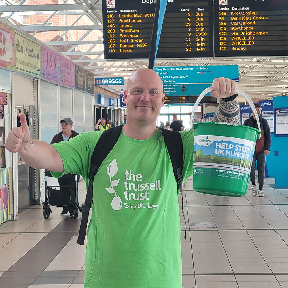 A fundraiser stands underneath a departure board in a bus station. They are wearing a green Trussell Trust t-shirt and backpack. They are smiling into the camera while holding a fundraising bucket in one hand and giving a thumbs-up with the other.
