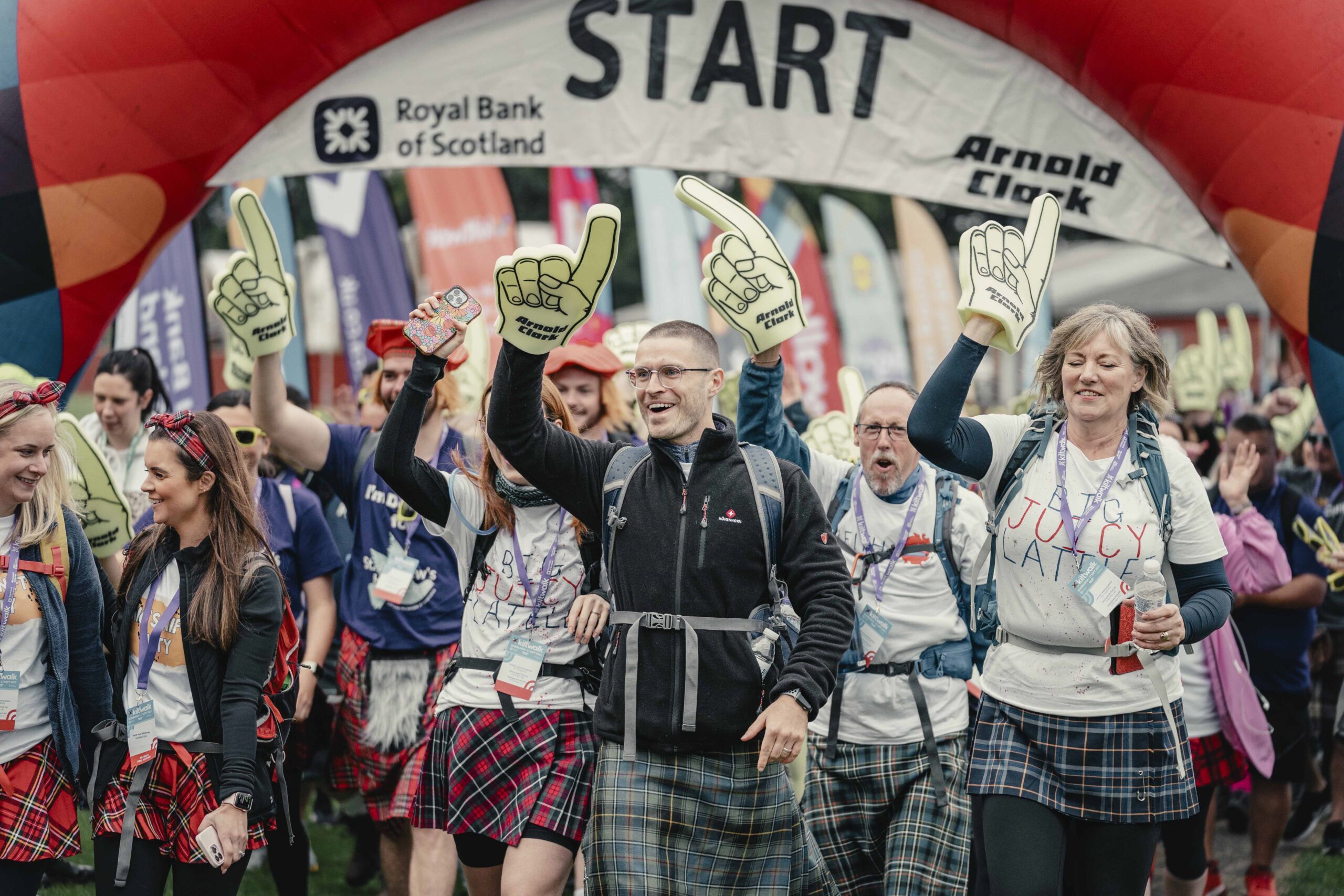 Participants in the Kiltwalk walk from the start line. All are wearing kilts, smiling and raising their hands in the air.