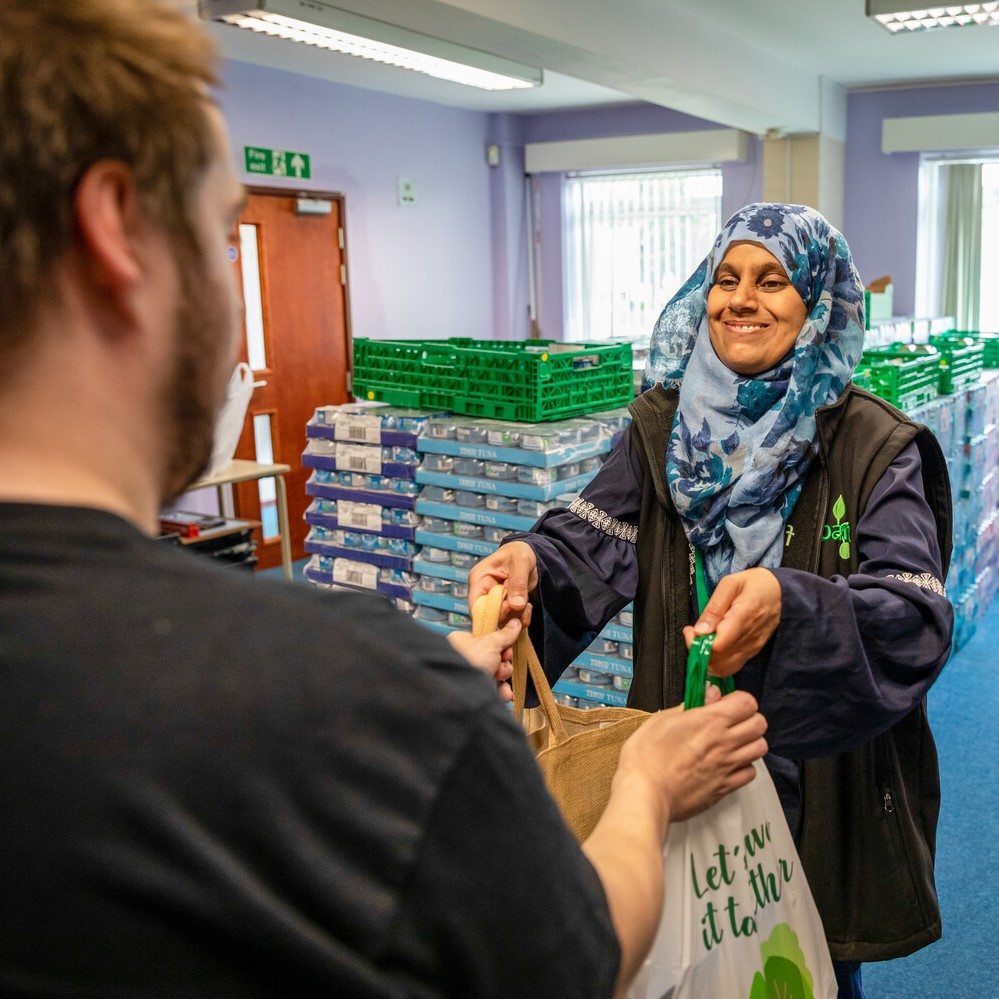 A food bank volunteer smiles and hands over bags of food to a visitor. They are wearing a blue patterned hijab and a black food bank top.